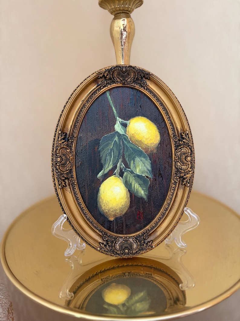 Pode incluir: Uma pintura a &oacute;leo de formato oval apresentando dois lim&otilde;es amarelo-brilhantes pendurados num caule com folhas verdes contra um fundo castanho escuro, inserida numa moldura dourada ornamentada com detalhes em relevo e base.