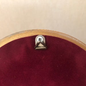 Pode incluir: Um objeto circular com um verso de veludo bord&ocirc; profundo e uma borda dourada. Um pequeno suporte triangular de metal para pendurar na parede est&aacute; fixado no centro superior da superf&iacute;cie de veludo com um parafuso de cor prateada.
