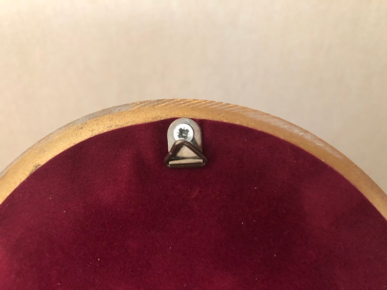 Pode incluir: Um objeto circular com um verso de veludo bord&ocirc; profundo e uma borda dourada. Um pequeno suporte triangular de metal para pendurar na parede est&aacute; fixado no centro superior da superf&iacute;cie de veludo com um parafuso de cor prateada.