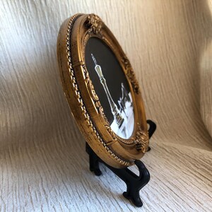 May include: An oval picture frame with a metallic gold finish and ornate, raised detailing along the border. The frame contains a black and white image of a tall, slender chess piece, specifically a king, set against a dark background.