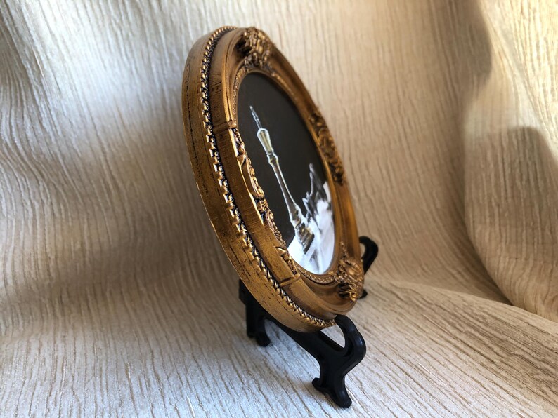 May include: An oval picture frame with a metallic gold finish and ornate, raised detailing along the border. The frame contains a black and white image of a tall, slender chess piece, specifically a king, set against a dark background.