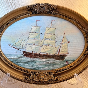 May include: An oval-shaped oil painting featuring a sailing ship with white sails on a blue sea under a light sky. The artwork is encased in an ornate, metallic gold-toned frame with intricate embossed floral and scrollwork detailing.