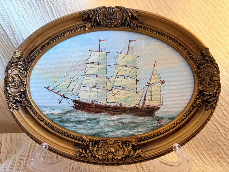 May include: An oval-shaped oil painting featuring a sailing ship with white sails on a blue sea under a light sky. The artwork is encased in an ornate, metallic gold-toned frame with intricate embossed floral and scrollwork detailing.