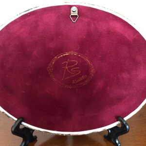 May include: An oval-shaped decorative plaque featuring a deep burgundy velvet backing. A gold-toned metal triangular hanging hook is attached at the top. In the center, a gold-colored circular emblem displays the initials RS and the text R.SoilArt.