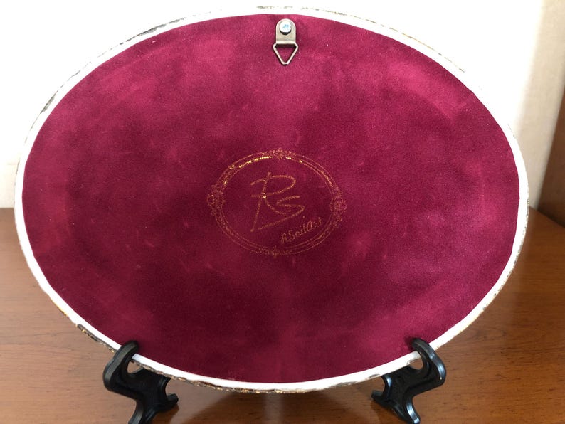May include: An oval-shaped decorative plaque featuring a deep burgundy velvet backing. A gold-toned metal triangular hanging hook is attached at the top. In the center, a gold-colored circular emblem displays the initials RS and the text R.SoilArt.