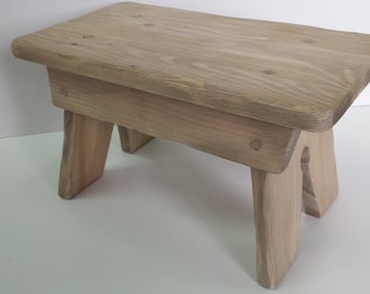 Peut inclure: Un tabouret en bois simple avec une finition naturelle. Le tabouret a un dessus rectangulaire et quatre pieds.