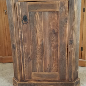 May include: A rustic wooden cabinet with a single door and a black metal ring pull handle. The cabinet has a dark brown finish and is made of reclaimed wood.