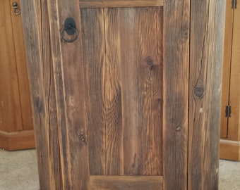 Può includere: Un armadio in legno rustico con un'unica porta e una maniglia ad anello in metallo nero. L'armadio ha una finitura marrone scuro ed è realizzato in legno riciclato.
