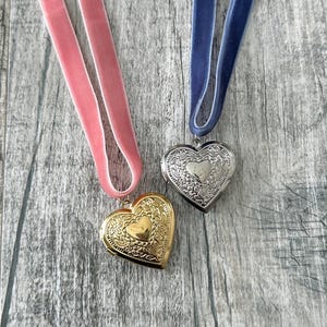 Puede incluir: Dos colgantes en forma de corazón en cintas de terciopelo. Un colgante es de oro con un diseño adornado, el otro es de plata con un diseño similar. Los colgantes están sobre un fondo de madera.