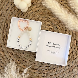Könnte beinhalten: Eine weiße Geschenkbox mit einem weißen und goldenen Perlenarmband, auf dem "Patente" steht, und einem goldenen herzförmigen Schlüsselanhänger. Die Box enthält auch eine weiße Karte mit der Aufschrift "Willst du meine Patenante sein? Tanja".