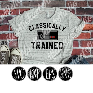 Könnte beinhalten: Ein graues Melange-T-Shirt mit dem Text "Classically Trained" und einer Schwarzweiß-Illustration eines Vintage-Videospiel-Controllers. Das Shirt liegt auf einem Ziegelhintergrund. Das Bild enthält auch SVG-, DXF-, EPS- und PNG-Dateityp-Symbole.