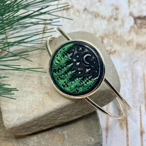 May include: A silver-toned bangle bracelet with a circular pendant. The pendant features a green forest design under a starry night sky. The bracelet is resting on two gray stones, with a sprig of greenery and red berries in the background.