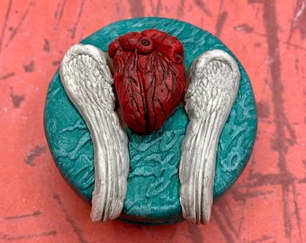 May include: A teal blue round trinket box with a red heart and silver wings on top.