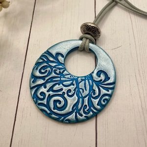 以下が含まれることがあります： ターコイズブルーのペンダントネックレス。シルバーの留め具と装飾的なビーズが付いています。ペンダントは、中央にくり抜かれた円形のある大きな楕円形で、シルバーのフローラルデザインが施されています。