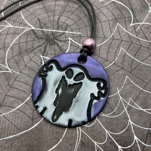 May include: A Halloween-themed pendant necklace featuring a round, purple and silver pendant with a black ghost design. The pendant is suspended from a black cord necklace with a purple bead accent. The background has a spiderweb design.