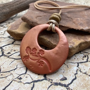 May include: A copper-colored necklace with a round pendant featuring a lizard design. The pendant has a large hole in the center and is attached to a beige cord with a wooden bead.