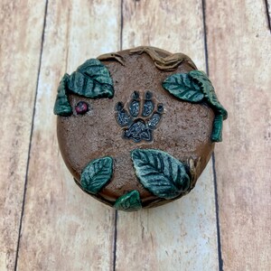 May include: A round, brown, ceramic trinket box with a paw print design and green leaves around the edges.