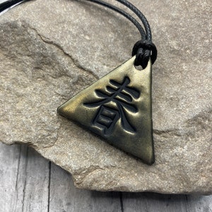 Puede incluir: Un collar con un colgante de triángulo dorado y un cordón negro. El colgante presenta el carácter japonés para "primavera" en negro.