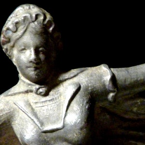 May include: A silver statue of a woman with her arms outstretched. She is wearing a long, flowing robe with a high collar.