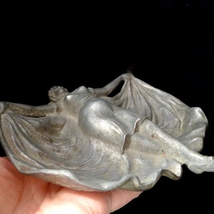 May include: A silver metal ashtray shaped like a reclining figure with flowing drapery. The figure is in a relaxed pose with arms outstretched and legs extended.