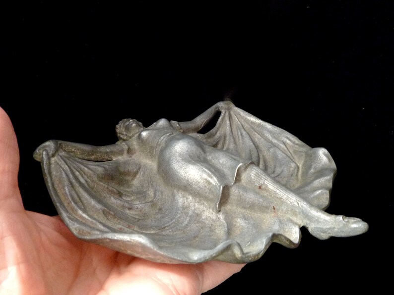 May include: A silver metal ashtray shaped like a reclining figure with flowing drapery. The figure is in a relaxed pose with arms outstretched and legs extended.
