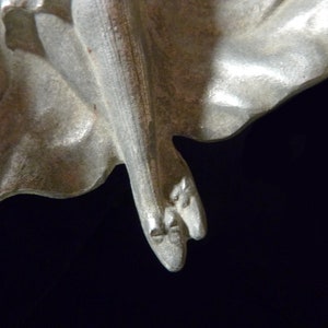 May include: A silver-toned metal sculpture of a woman's legs and feet. The sculpture is detailed with folds of fabric and the woman's toes are visible.