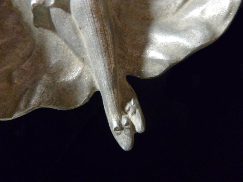 May include: A silver-toned metal sculpture of a woman's legs and feet. The sculpture is detailed with folds of fabric and the woman's toes are visible.