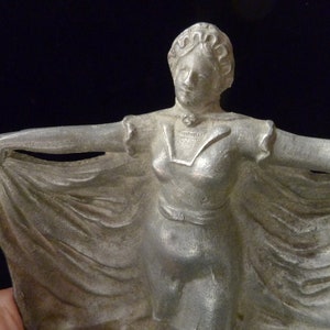 May include: A silver metal figurine of a woman in a flowing dress. The woman is holding the edge of her dress with both hands and is looking to the right. The figurine is a vintage style and has a detailed design.