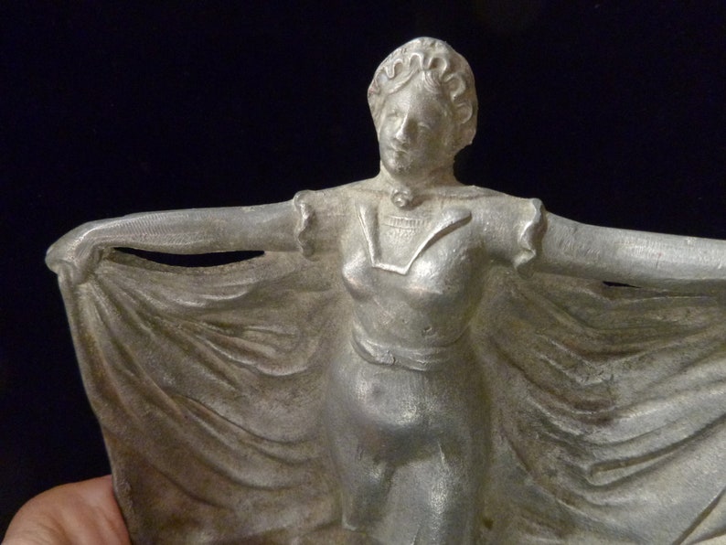 May include: A silver metal figurine of a woman in a flowing dress. The woman is holding the edge of her dress with both hands and is looking to the right. The figurine is a vintage style and has a detailed design.
