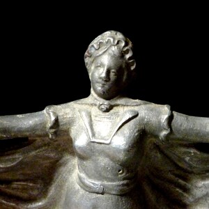 May include: A silver statue of a woman wearing a long dress and a crown. She is standing with her arms outstretched.