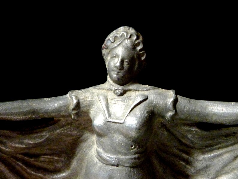 May include: A silver statue of a woman wearing a long dress and a crown. She is standing with her arms outstretched.