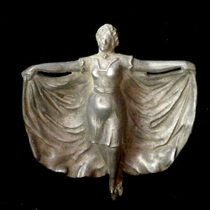 May include: A silver-toned metal sculpture of a woman in a flowing dress. The woman is standing with her arms outstretched and her dress is billowing out behind her.