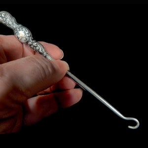May include: A silver button hook with an ornate handle. The hook is curved and has a pointed end.