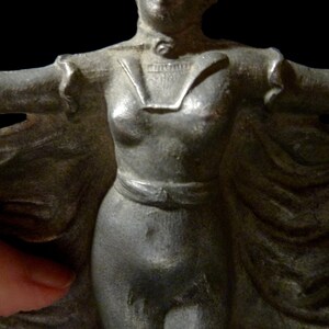 May include: A silver-colored metal figurine of a woman wearing a long flowing cape. The woman is standing with her arms outstretched and her head tilted slightly to the side. The figurine is detailed with a textured surface and a belt around the waist.