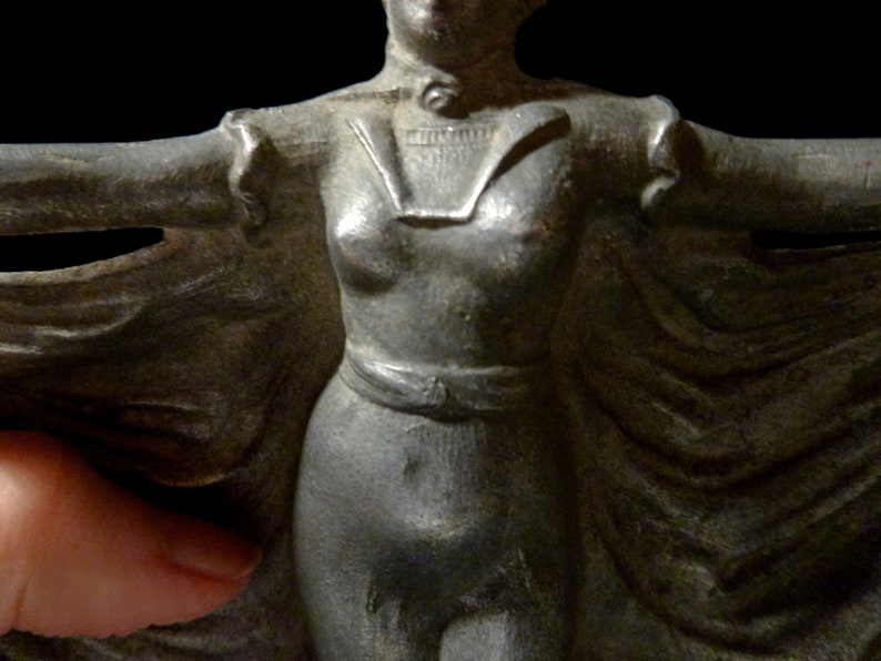 May include: A silver-colored metal figurine of a woman wearing a long flowing cape. The woman is standing with her arms outstretched and her head tilted slightly to the side. The figurine is detailed with a textured surface and a belt around the waist.