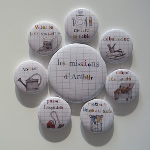 May include: Nine colourful round buttons with illustrations and text in French. The buttons feature tasks like "Vider le lave-vaisselle" (empty the dishwasher), "mettre la table" (set the table), "d&eacute;barrasser" (clear the table), "arroser" (water), "les missions d'Arthur" (Arthur's missions), "ranger les jouets" (put away the toys), "passer l'aspirateur" (vacuum), and "mettre le linge au sale" (put the laundry in the laundry basket).
