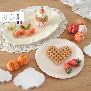 Peut inclure: Une collection d'aliments en crochet, dont un cupcake, une gaufre, des fraises, des macarons et des oranges. Les articles sont disposés sur des assiettes blanches et roses et une surface en bois.