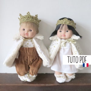May include: Two dolls wearing white capes with gold star patterns and gold crowns. The doll on the left is wearing brown corduroy trousers and the doll on the right is wearing a white dress. The text "Tuto PDF" is visible in the image.