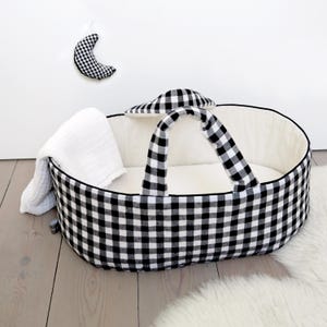 Può includere: Una culla ovale per neonati con motivo a quadretti vichy bianchi e neri, dotata di due manici abbinati e rivestimento interno color crema. Una coperta in mussola bianca è drappeggiata sul lato e un giocattolo a forma di luna è appeso.