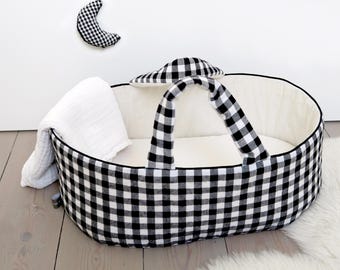Op de afbeelding: Een ovale babywieg met een zwart-wit vichy-ruitpatroon, voorzien van twee bijpassende handvatten en een crèmekleurige binnenvoering. Een witte mousseline deken hangt over de zijkant en er hangt een speelgoedje in de vorm van een halve maan.
