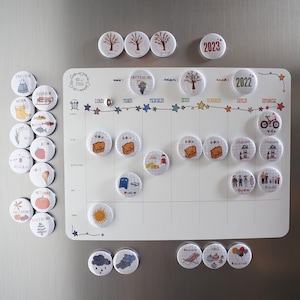 Peut inclure: Un calendrier magnétique blanc avec des aimants illustrés colorés pour chaque mois de l'année. Le calendrier a une grille avec les jours de la semaine étiquetés en français. Les aimants présentent des images d'activités, de fêtes et de saisons.