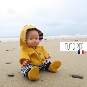 May include: A yellow raincoat with a hood and a striped lining, worn by a doll sitting on a sandy beach. The doll has blue eyes and is wearing striped pants. The text "TUTO PDF" is visible in the image.