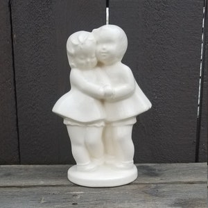 May include: White ceramic figurine of two girls holding hands and hugging each other. The girls are wearing dresses and have short hair.