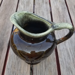 May include: A small, brown ceramic pitcher with a green glaze. The pitcher has a rounded body and a handle.