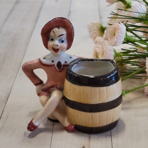 May include: A ceramic figurine of a woman with a red hat and dress sitting next to a small brown and black barrel. The figurine is painted with details and has a whimsical expression.