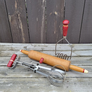 May include: A set of three vintage kitchen tools: a red and white rolling pin, a red and white potato masher, and a silver whisk with a red handle.