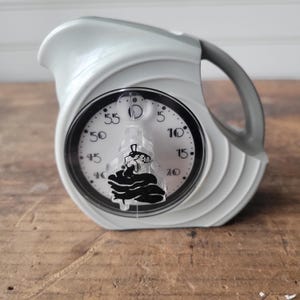 May include: A white and grey kitchen timer in the shape of a jug. The timer features a black and white clock face with a black graphic. The handle is grey. It is on a wooden surface.