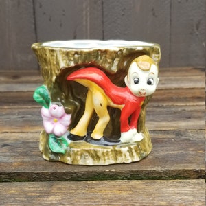 May include: A ceramic planter shaped like a tree stump with a small, yellow deer figurine wearing a red cape. The deer is peeking out from behind the tree stump. There is a pink flower with green leaves on the side of the planter.