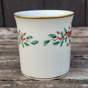 May include: White ceramic mug with a gold rim and a festive holly and berry design. The mug has a white background with green holly leaves, red berries, and gold stars.