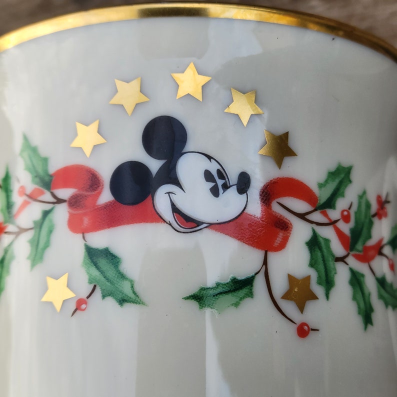May include: White ceramic mug with a gold rim featuring a cartoon image of Mickey Mouse wearing a red ribbon with holly and gold stars around him.